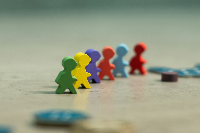A photo of severl person-shaped meeple in many colors walking on a tan board