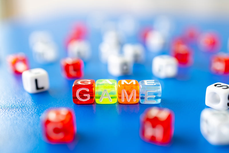 A photograph of an assortment of dice and beads on a blue tabletop. There are four beads in the center, lined up so that their letters spell out "GAME"