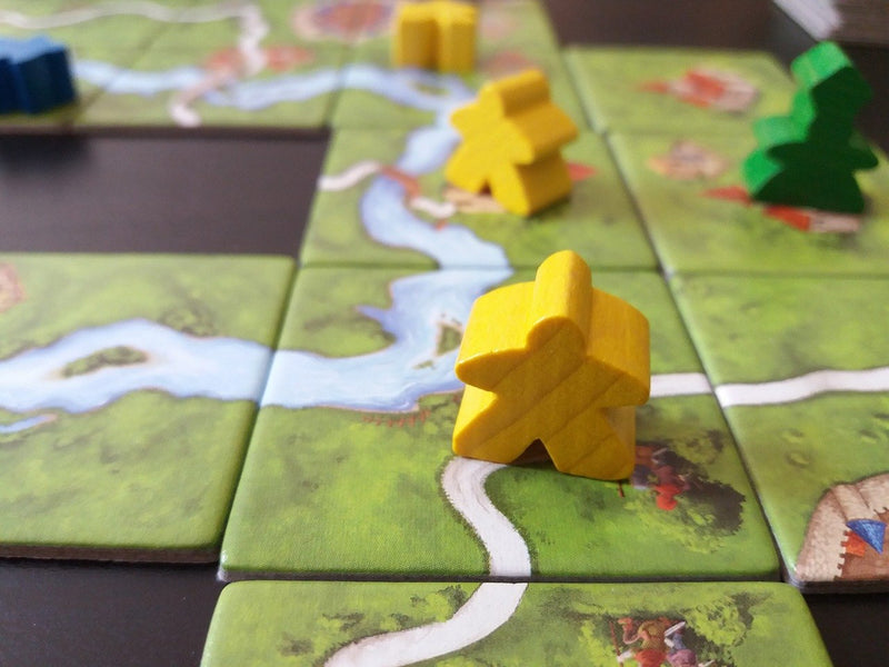 A photo of several meeples, focusing on a front, central yellow meeple, standing on several connected squares with forests, roads, and rivers printed on them