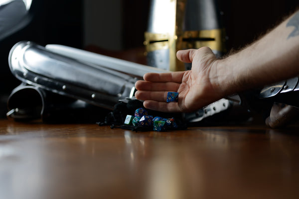 A photograph of a person's hand rolling a die into a small pile of other dice. Behind this is a helmet and gauntlets for a suit of medieval armor