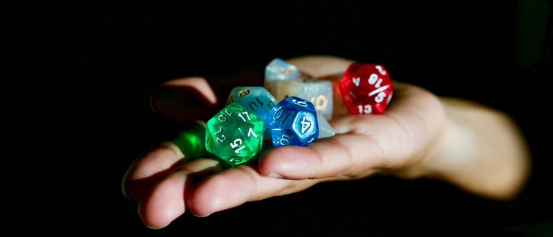 Photo of a hand held out flat, on which are several transparent dice in different colors, with white numbers. The hand emerges out of a completely black background. 