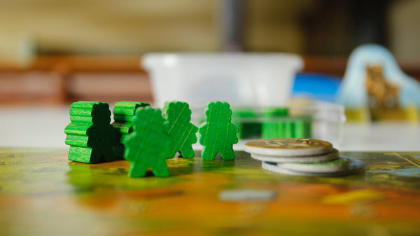 A photograph of a boardgame set up on a table. Several small green meeples are stood on the middle of a board, with a small stack of round cardboard tokens and a tupperware container near them