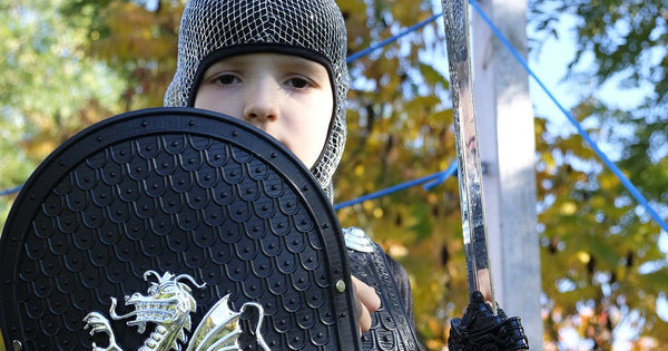 A photo of a young child wearing a knight costume, as though he is LARPing. He holds a plastic sword and shield, and ears a chainmaille coiffe