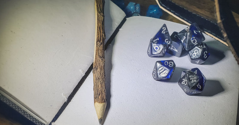 A photo of an open leather journal, with a set of blue and silver dice and a wooden carved pencil sitting on a blank page.