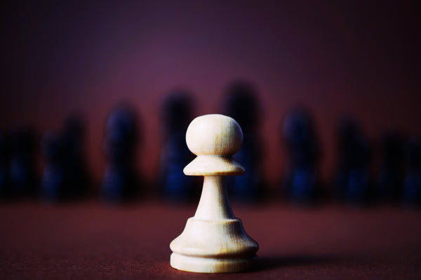 A photograph of a single white pawn in front of several black chesspeices. 