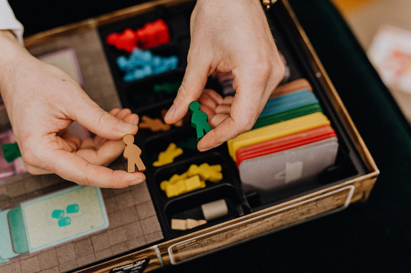 A photo of a pair of hands holding yellow and green meeples above a boardgame box, in which there are cards, various smaller tokens, and black dividers