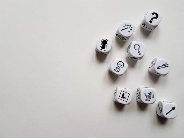 A photo of 9 dice with various images on each side, such as a magnifying glass, a keyhole, a question mark, an arrow, and a lightbulb, against a white background
